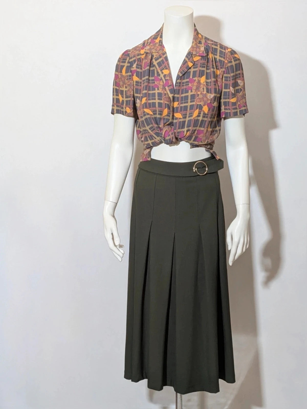 Mangano green military skirt suit with elegant front pleats and metallic ring detail.